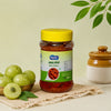 A jar of Amla Pickle (sour orange) with a yellow lid and green label, styled on a beige cloth with whole and sliced green amla fruits, a small brown ceramic jar, and a leafy branch against a light green background.