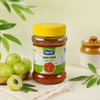 A jar of Amila Redde (amaranthum) spread by Shiv Kumar, featuring a bright yellow lid and a green and white label, styled with green amlas and leaves on a light beige surface.