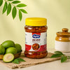 A jar of mango pickle with a bright yellow lid and a red-and-white label, styled on a woven mat with green mangoes, leaves, and a ceramic container against a pale yellow background.