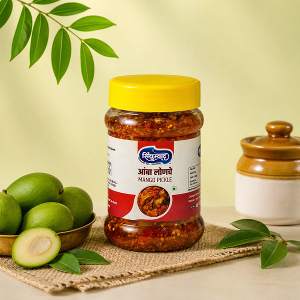 A jar of mango pickle with a bright yellow lid and a red-and-white label, styled on a woven mat with green mangoes, leaves, and a ceramic container against a pale yellow background.
