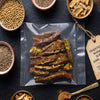 A clear plastic bag containing dried, sliced chili peppers or hot sauce pieces, arranged on a dark surface with bowls of spices and seeds around it. A paper tag reads 'Sindhuswad Foods'..