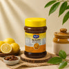 A jar of Sindhuswad Foods Sweet Lemon Pickle is displayed on a rustic wooden slice, surrounded by fresh lemons, a small bowl of pickle, and a ceramic pot against a warm beige background with soft leaf shadows.